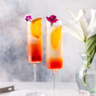 Aperol spritzes on a white background with a bouquet in the foreground of the shot. Each glass is decorated with small, edible purple flowers.