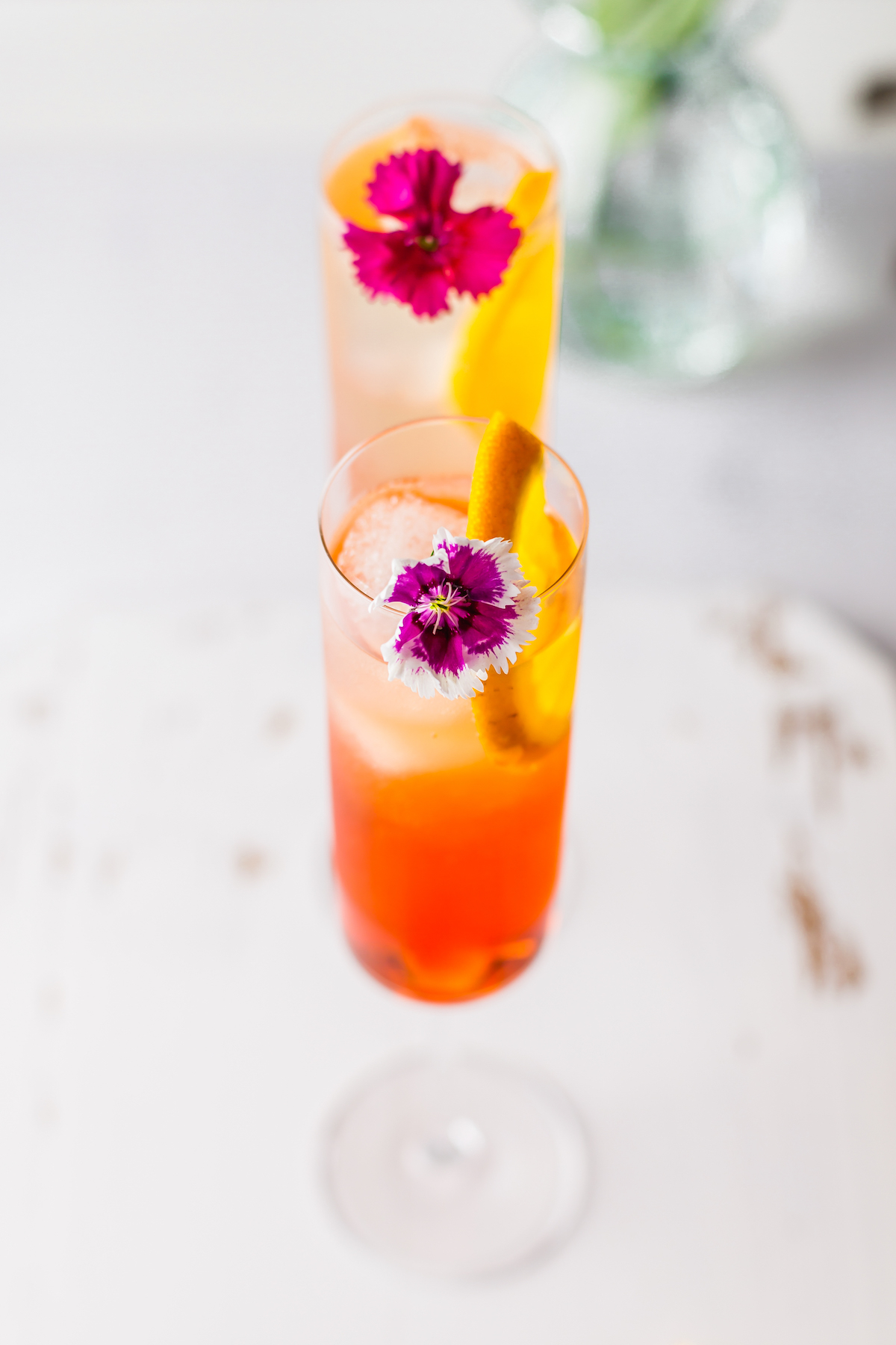 An overhead shot of two cocktails, garnished with edible flowers and orange slices, on a white table.