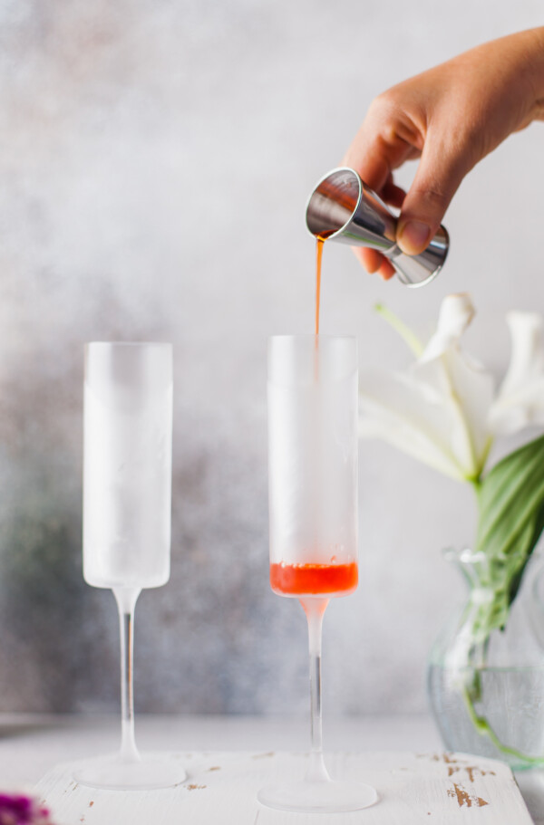 Aperol liqueur being poured into a glass against a white and gray background. A bouquet of white flowers is in the background of the shot.
