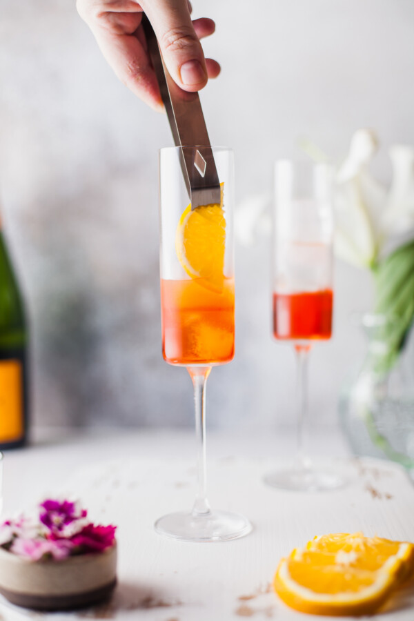 An orange is being placed inside the cocktail glass, against the side, on top of the ice and liqueur.