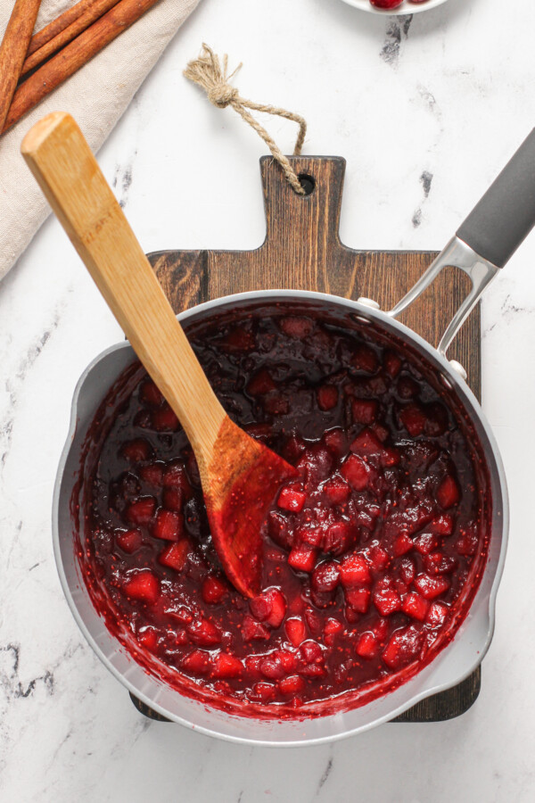 A saucepan with cooked apple cranberry sauce and a wooden spoon.