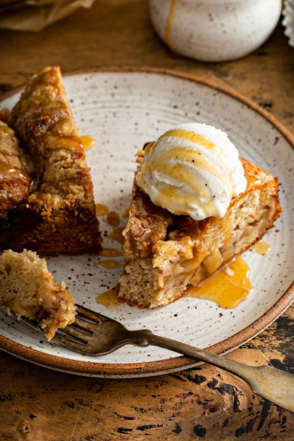 A slice of Apple Streusel Cake on a white plate with caramel on top and a fork taking a bite out of the cake.