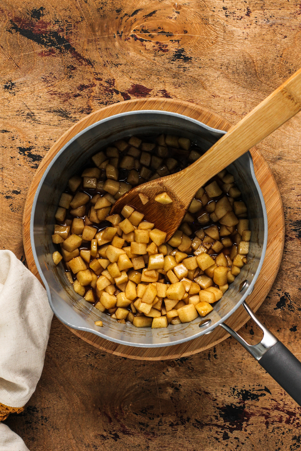 Diced apples in syrup in a metal sauce pan on a wooden trivet with a wooden spoon.