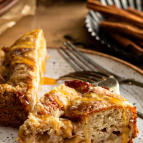 A slice of Apple Streusel Cake on a white plate with caramel drizzled on top with a fork in the back ground.