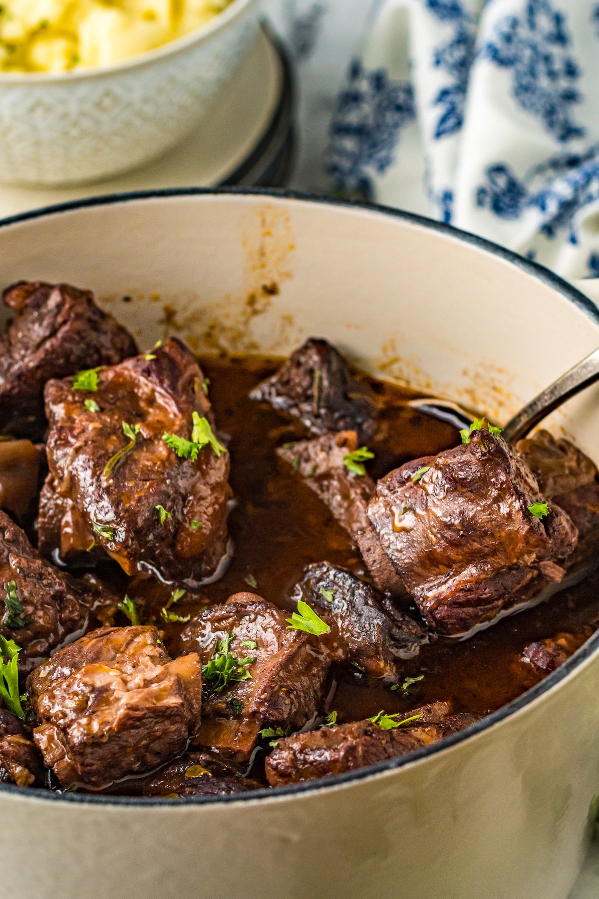Braised short ribs in a Dutch oven with rich brown sauce. A spoon is lifting one piece of the short ribs out of the sauce.