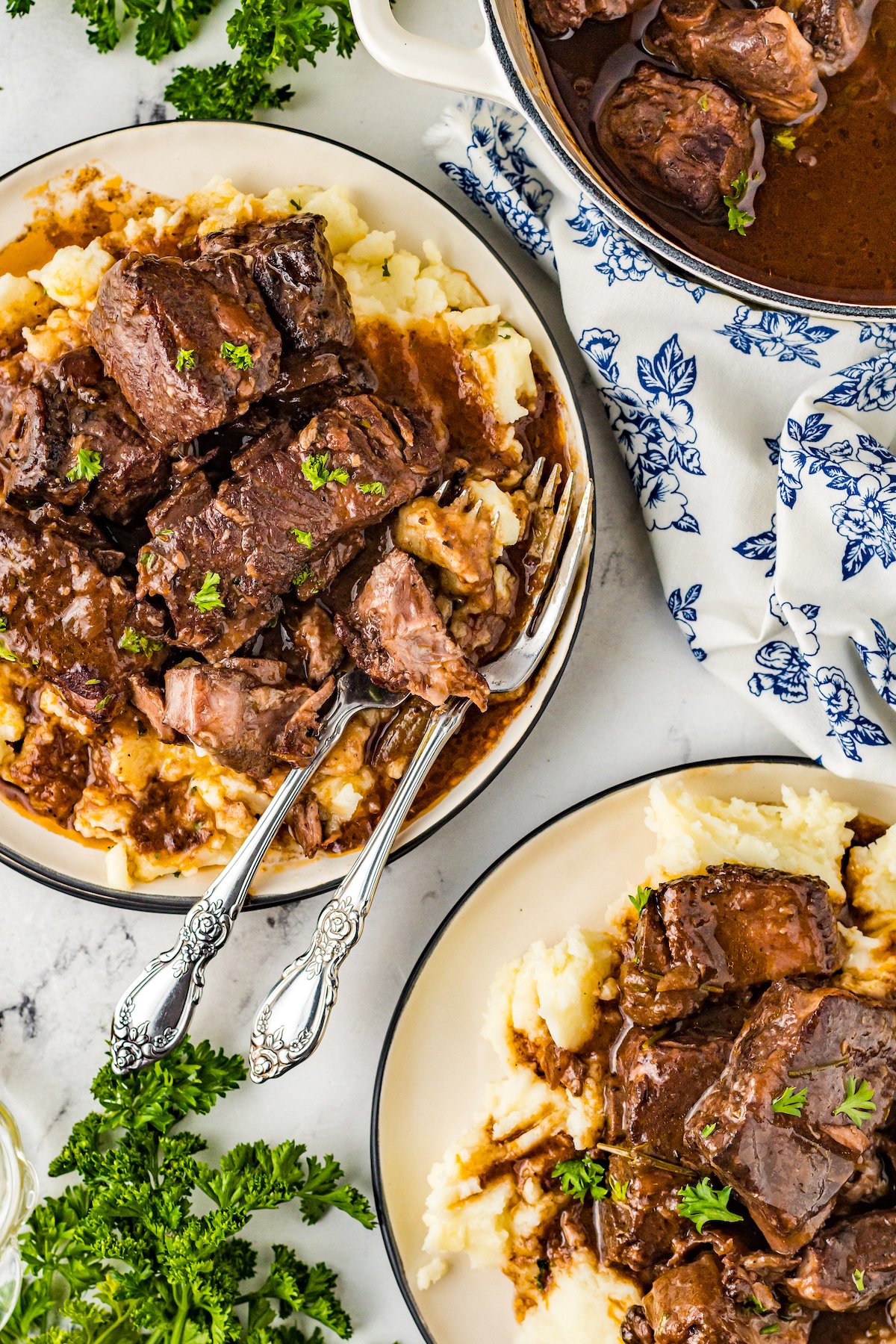 An overhead shot of two plates of short ribs, each on a bed of mashed potatoes. One of the plates also holds utensils. A blue-and-white napkin and the Dutch oven are partially visible in the shot, as is a bunch of fresh parsley.