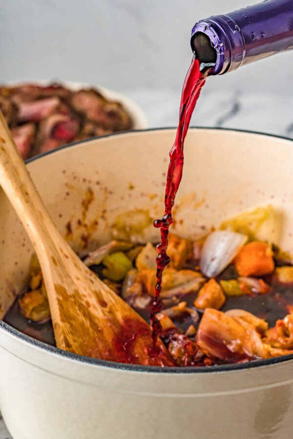 Red wine being poured into a Dutch oven with beef, vegetables, and broth inside.