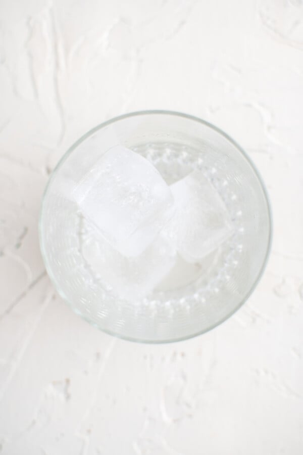 An overhead shot of a glass with several ice cubes at the bottom on a white work surface.