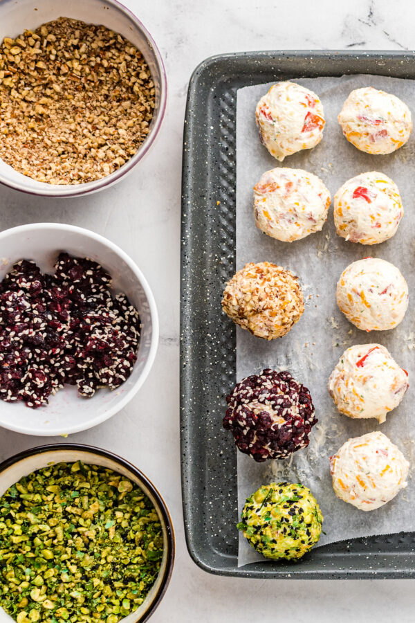 Three bowls, each holding a chopped topping, sit on a white surface next to a baking sheet lined with parchment. Cheese balls (topped and untopped) are lined up on the baking sheet.
