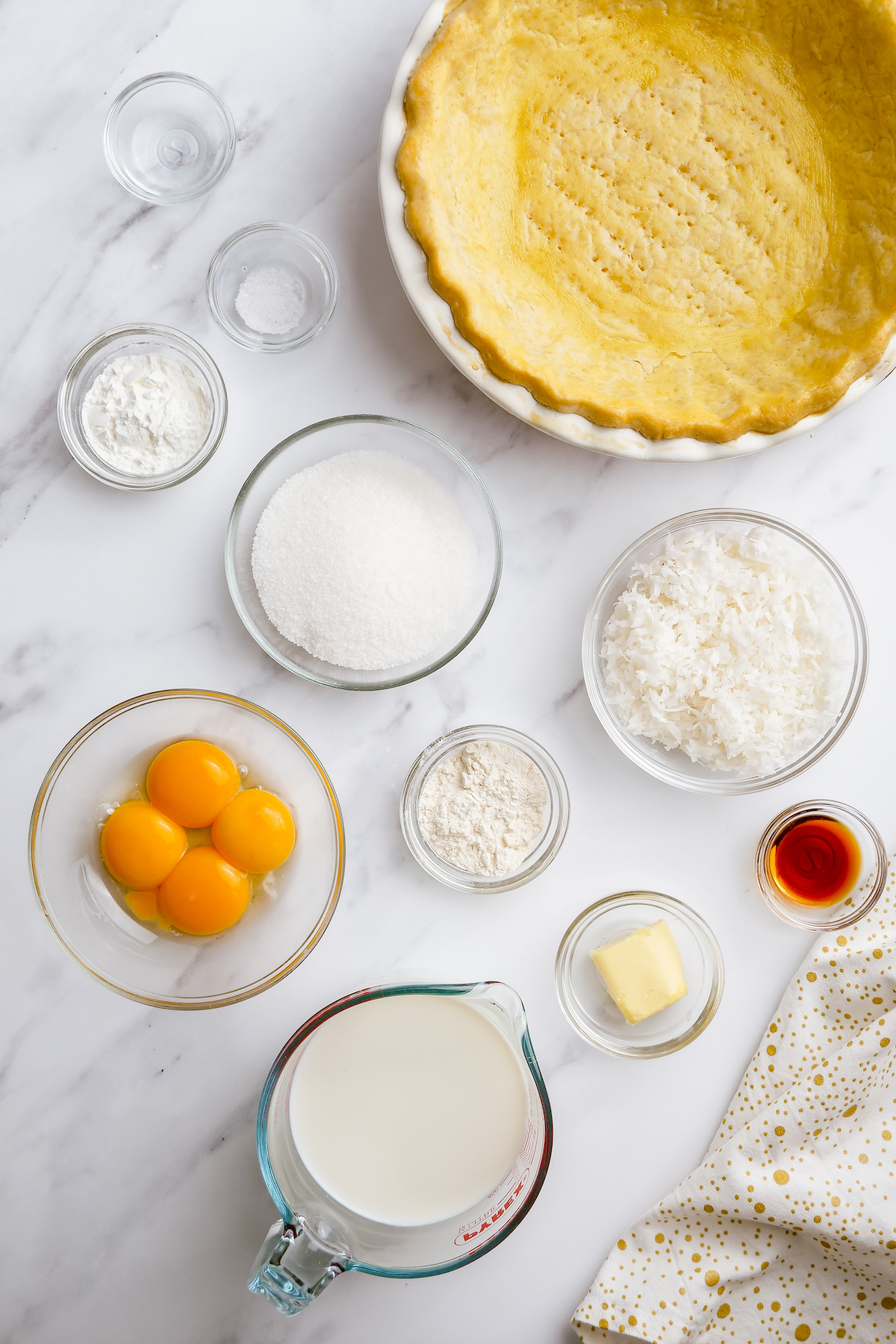 Clockwise from top: a baked pie crust, a bowl of sweetened shredded coconut, vanilla extract, butter, whole milk, egg yolks, flour, sugar, cornstarch, and coconut extract.