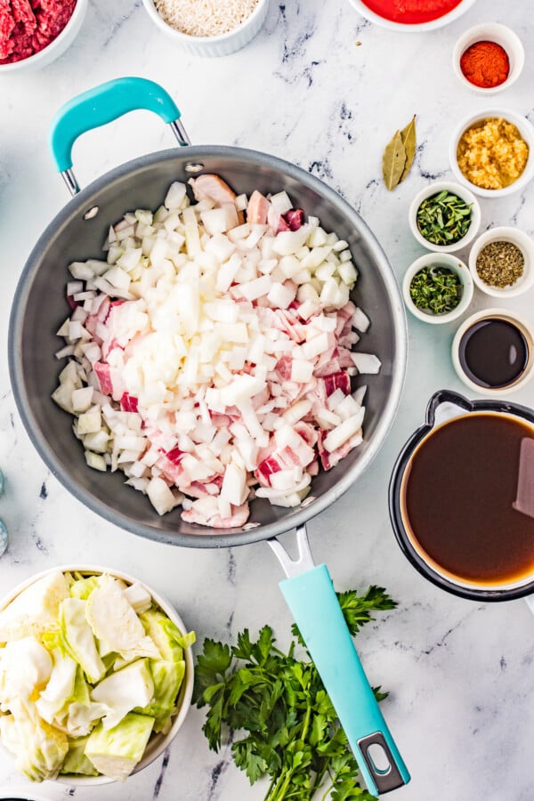 Bacon and onions in a skillet. Several small cups of dried herbs are nearby, along with a jumbo measuring cup of broth .