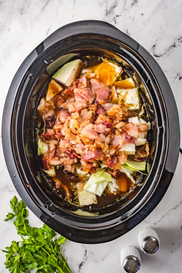 Overhead shot of an oval-shaped Crockpot insert, with layers of raw cabbage, canned tomatoes, bacon, onions, and other soup ingredients inside.