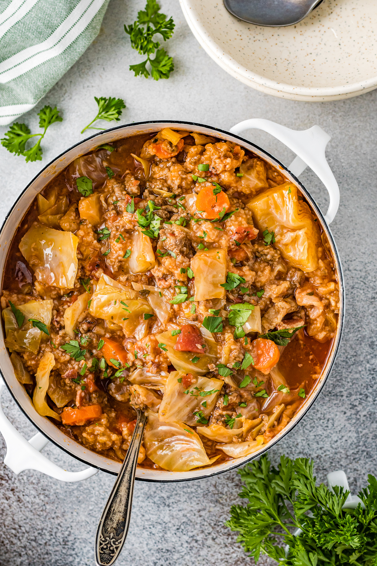 Overhead shot of a pot of cabbage roll soup, filled with vegetables, ground beef, and herbs in a tomato-based broth. A silver serving utensil is in the pot. Fresh herbs and a white bowl are nearby on the tabletop.