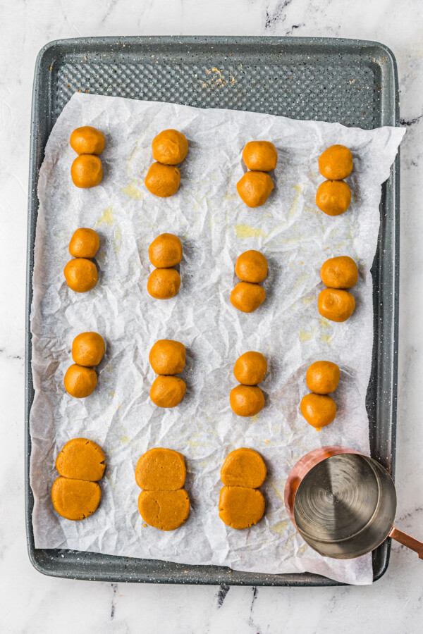 A cookie sheet lined with parchment. Rows of cookie dough ball pairs are on the cookie sheet. Four have been flattened and one has a metal measuring cup on top of it, flattening it.