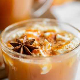 Image of pumpkin apple cider in a glass mug with whip cream, caramel, cinnamon and a star anise on top.