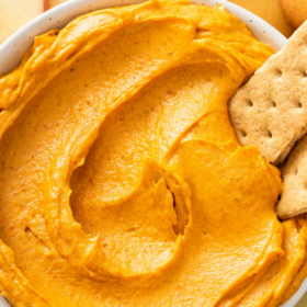 Up close image of pumpkin dip in a white bowl with two graham crackers dipped in it and fruit around the bowl.