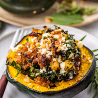 A rustic stuffed squash half filled with sausage, kale, and apples, topped with feta and nuts. In the background are two more stuffed squash on a platter.