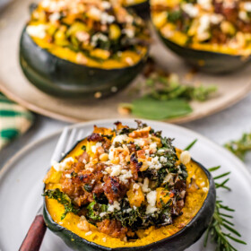 A serving of cooked stuffed acorn squash, with a platter of stuffed acorn squash in the background. Fresh herbs decorate the plates and a fork lies nearby.