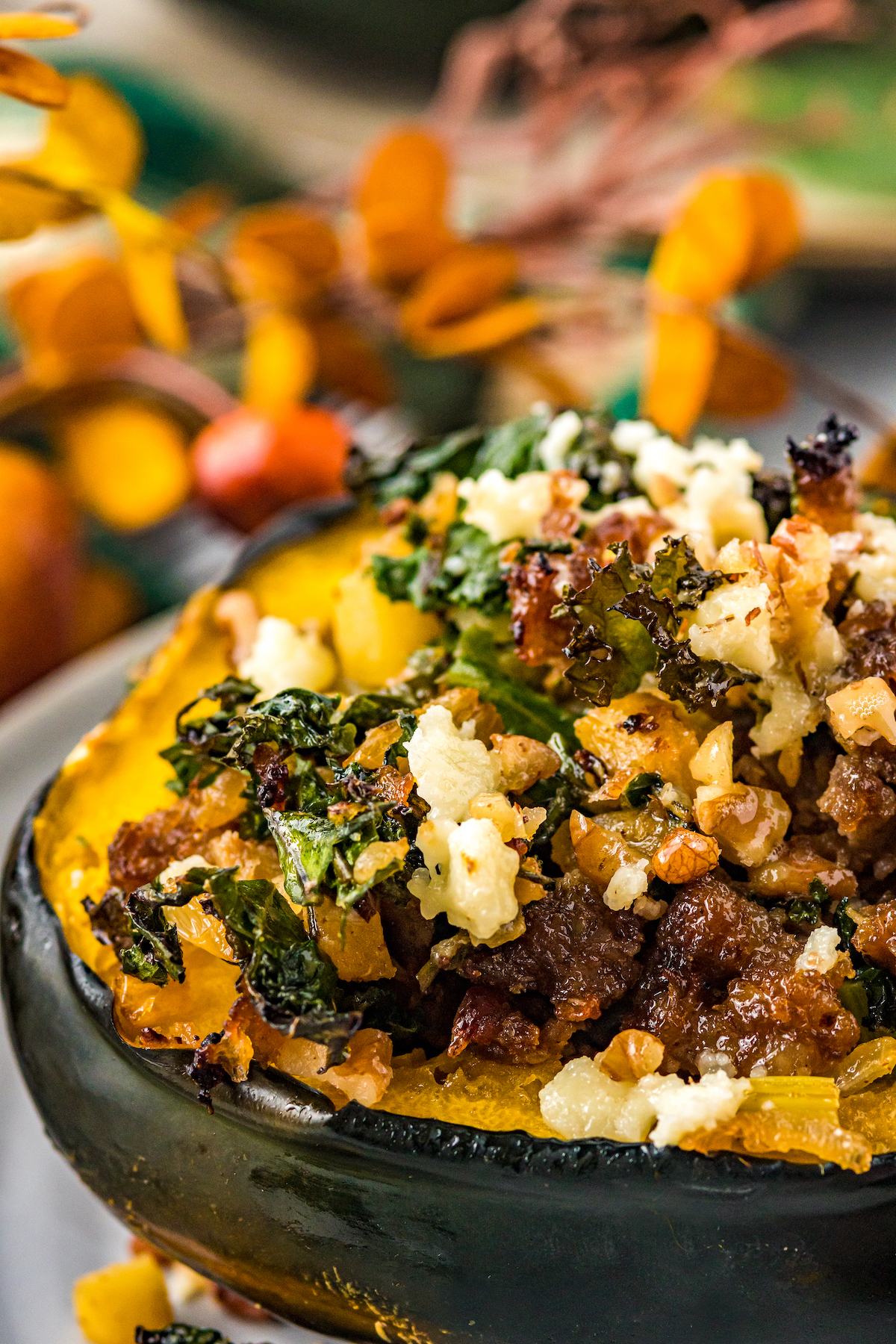 A close-up shot of an acorn squash stuffed with sausage, apples, kale, and more, topped with feta and nuts. Autumn decorations are in the background of the picture.