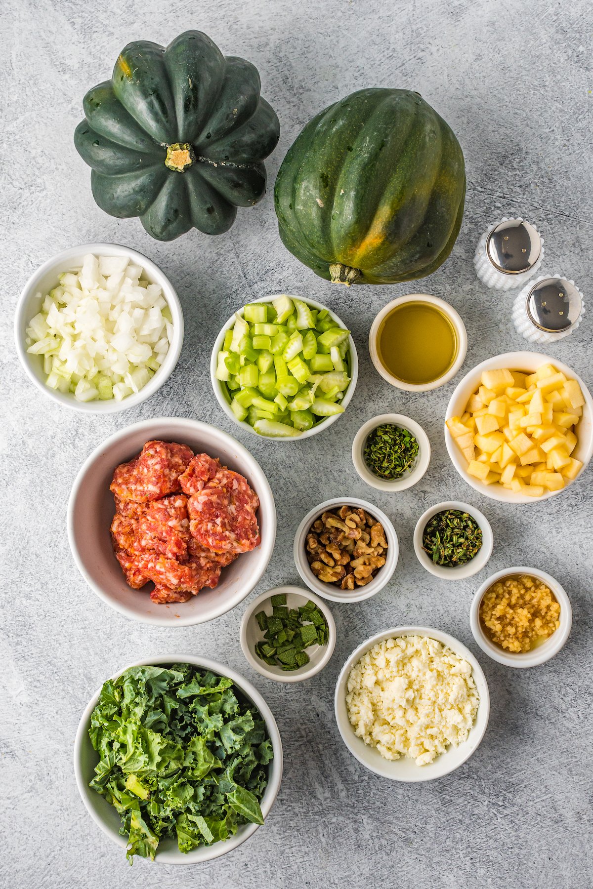 The ingredients for sausage stuffed squash: Whole acorn squash, salt, pepper, chopped apple. olive oil, minced herbs, garlic, feta cheese, kale, ground sausage, walnuts, celery, and onions.