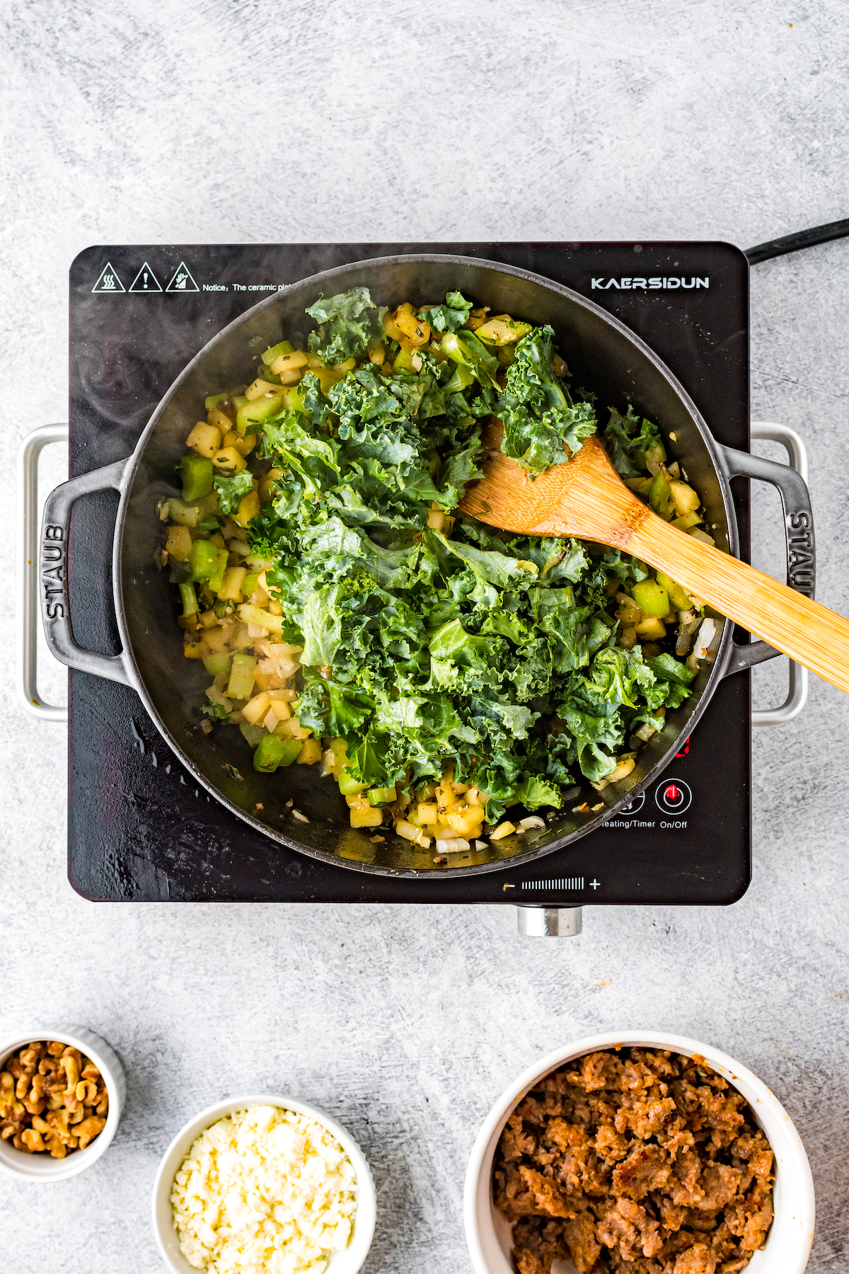 A skillet with apples, kale, onions, and other sausage filling ingredients. Cooked sausage in a bowl, as well as feta and chopped nuts, are nearby on the tabletop.