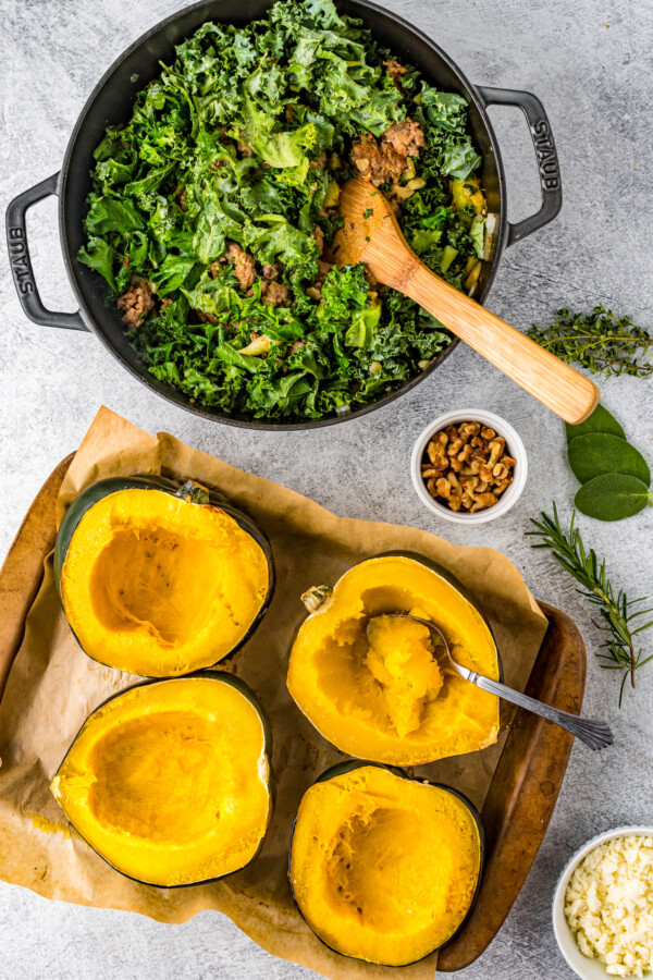Four roasted squash halves sit on a wooden tray, next to a cast-iron pot of sausage filling mixture.