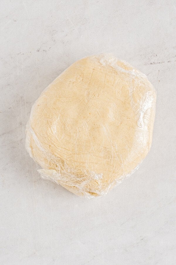 A disc of pale, unbaked sugar cookie dough, wrapped in plastic, on a white work surface.