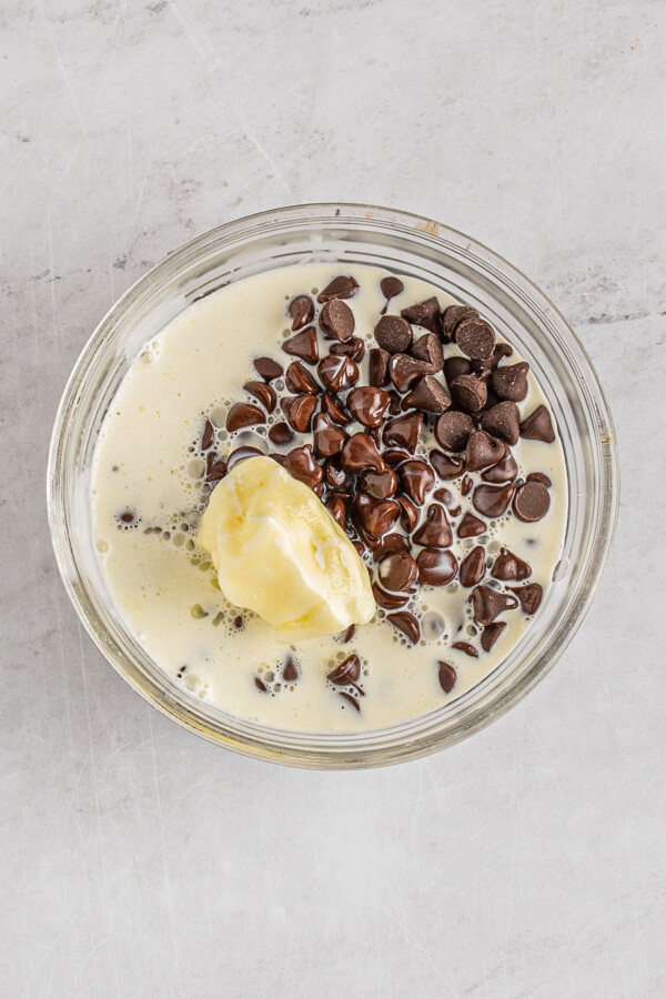 Chocolate chips melting in a clear glass bowl of hot cream, with a knob of butter in the bowl.