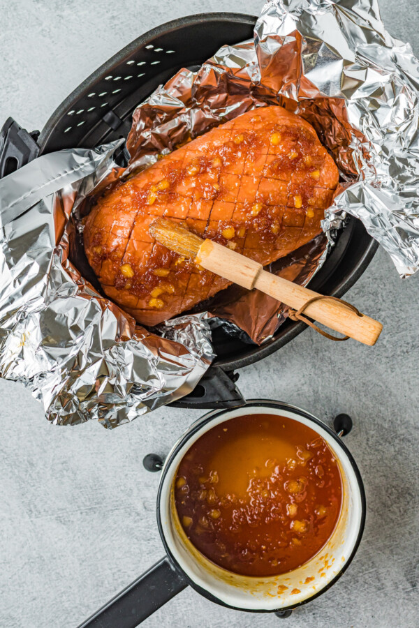 A ham in foil in an air fryer basket having sauce put on top before cooking.