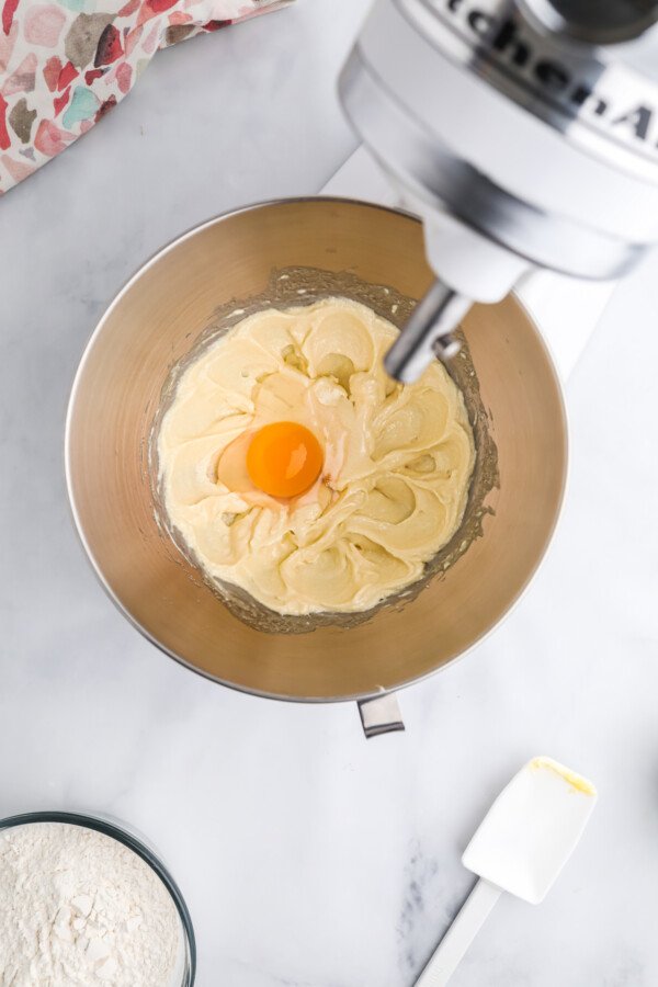 An overhead shot of the bowl of a stand mixer, with creamed butter and sugar inside, and a raw egg cracked into the bowl.