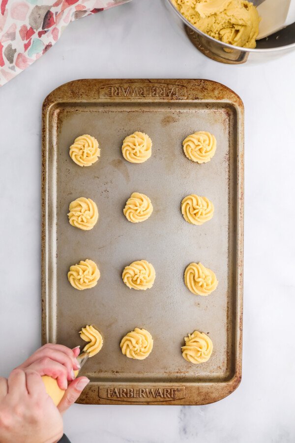 An overhead shot of a cookie sheet with twelve piped swirls of cookie dough on it. The twelfth swirl is being piped.