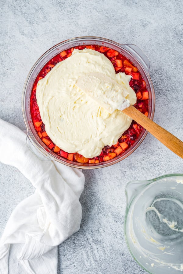 A bowl of set raspberry jello with fruit is being spread with a layer of whipped topping.