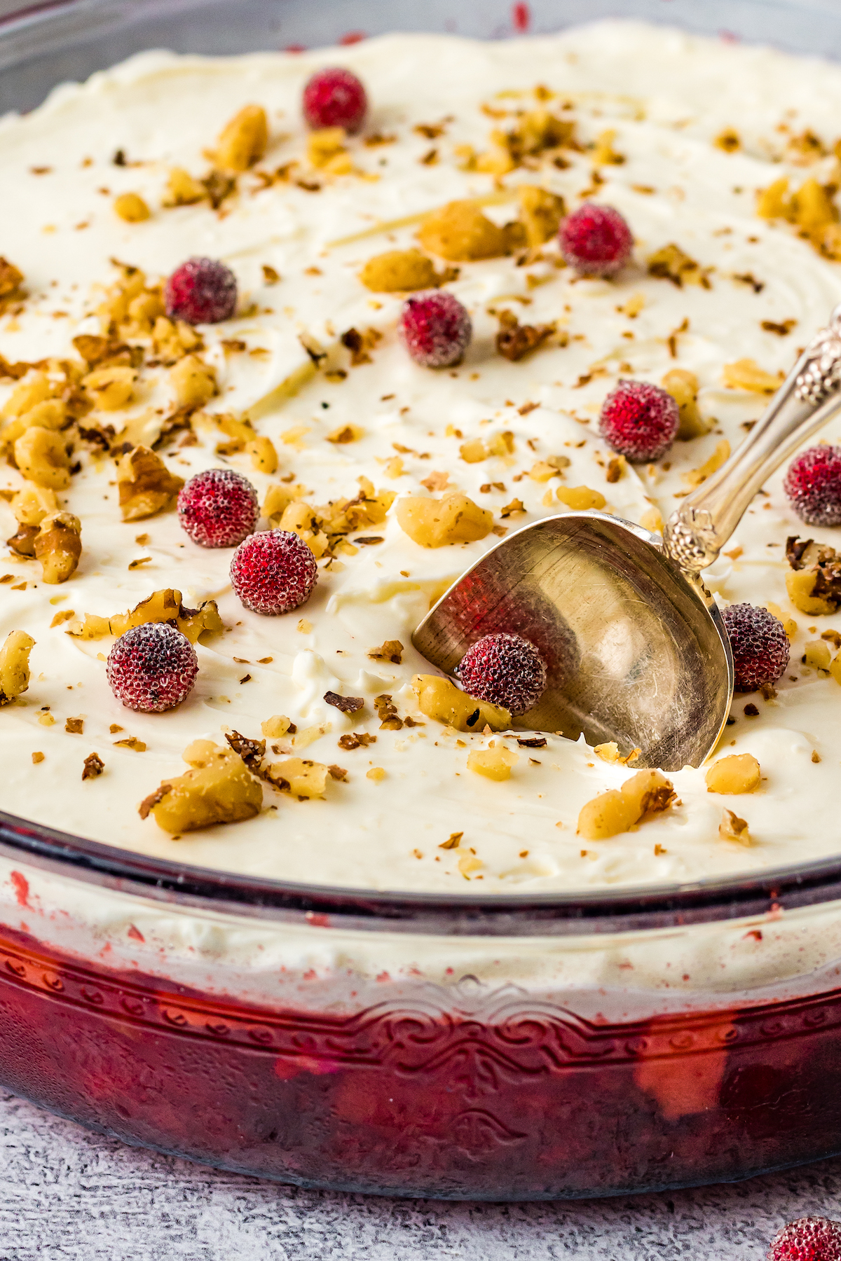 A glass bowl of jello salad with a red, fruit-filled bottom layer and creamy white top layer. The top is sprinkled with chopped nuts and sugared cranberries. A golden spoon is stuck into the creamy layer.