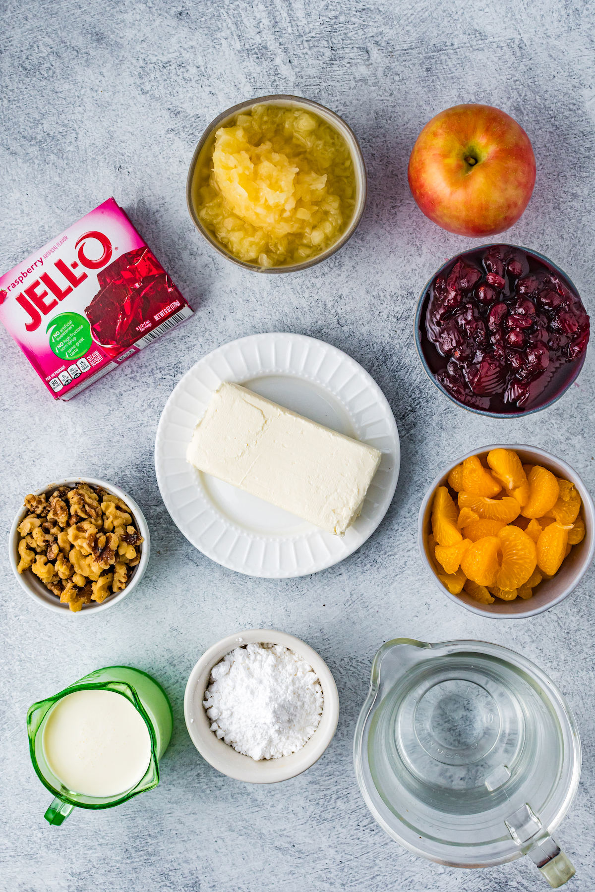 Clockwise from top: Crushed pineapple in juice, an apple, a bowl of cranberry sauce, a dish of mandarin segments, a measuring cup of water, a ramekin of powdered sugar, a container of cream, and a dish of chopped nuts. Center: a block of cream cheese.