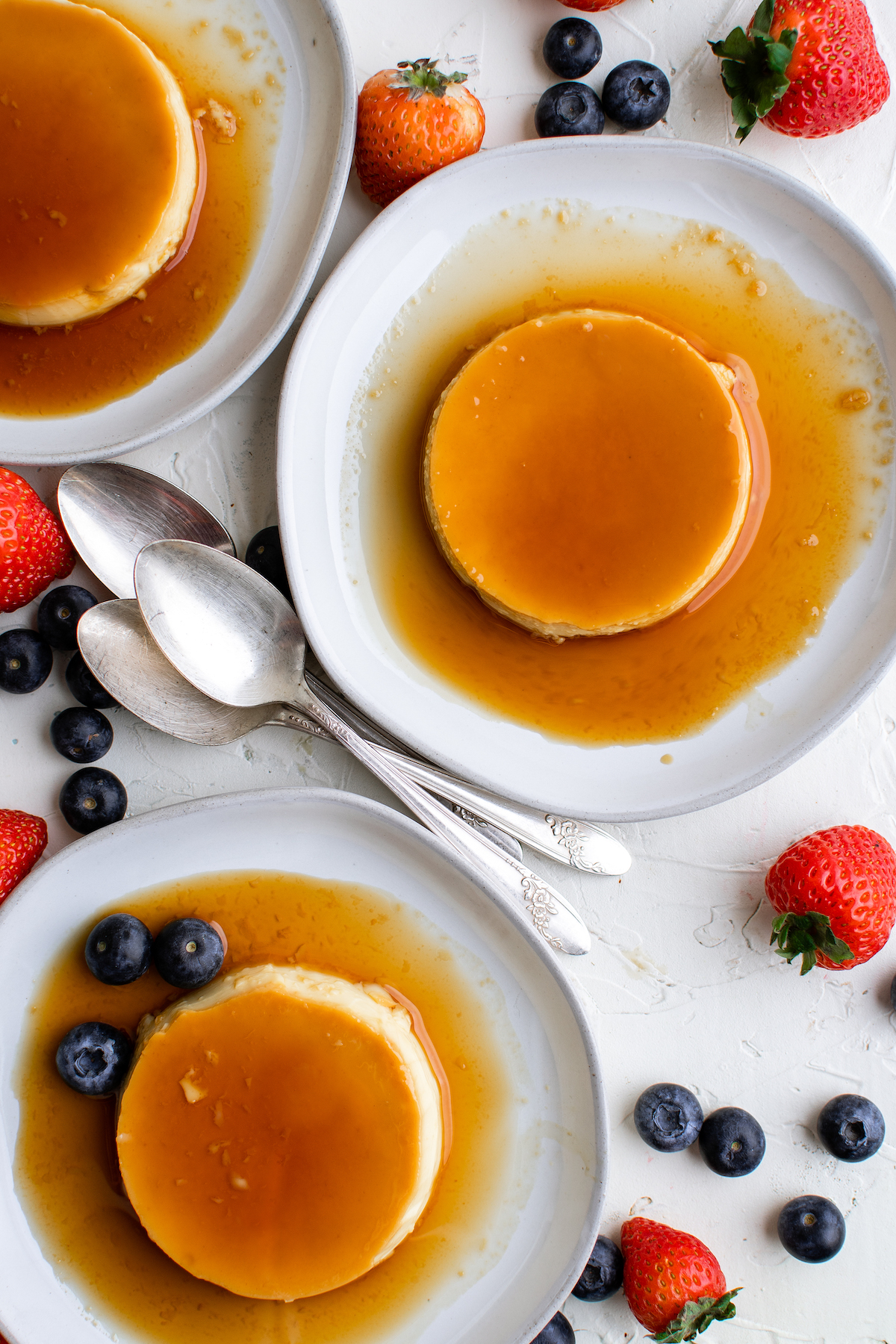Three individual crème caramel servings on saucers, on a white table scattered with strawberries and blueberries. Three spoons are arranged between the saucers.