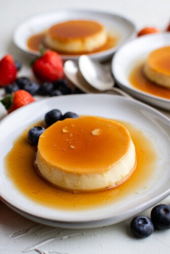 Three unmolded portions of crème caramel on saucers, with spoons and berries on the table nearby.