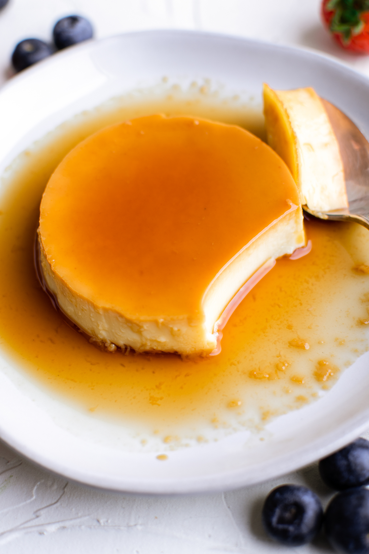 A serving of crème caramel with the sauce pooling on the plate. A bite-sized portion has been cut from the serving, and sits nearby resting on a spoon.