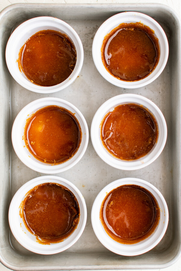 Six ramekins in a baking pan. Each ramekin has a small amount of cooled caramel in the bottom.