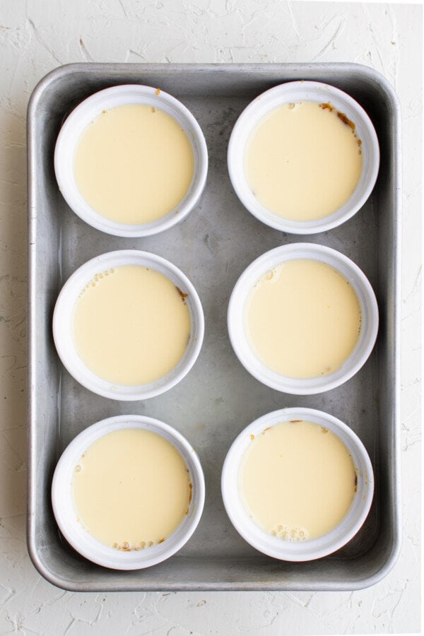 Six ramekins in a baking pan. Each ramekin is full of unbaked egg custard mixture.