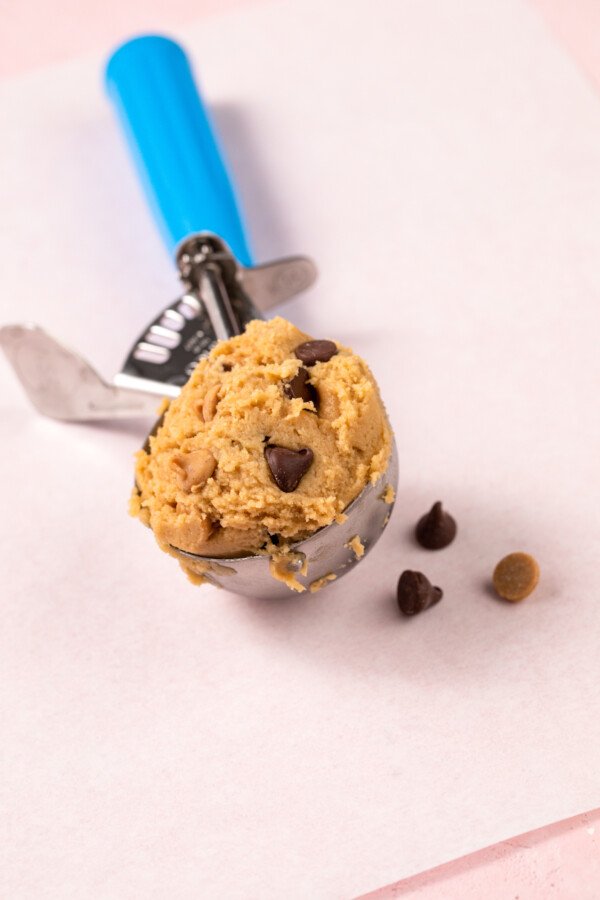 An ice cream scoop with a blue handle, filled with cookie dough. Two chocolate chips and one peanut butter chip are nearby on the work surface.