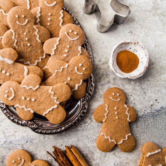 Gingerbread cookies are fanned out on an oval tray. On the table is a rustic dish of ground cinnamon, a cookie cutter, a completed gingerbread man, and some cinnamon sticks.