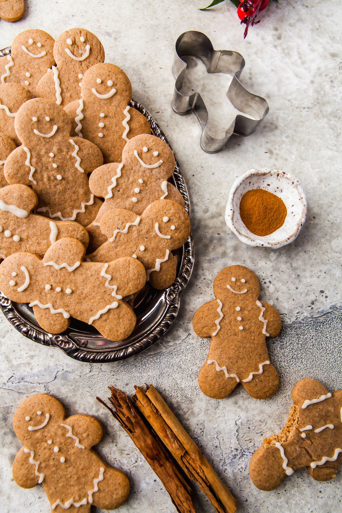 Gingerbread cookies are fanned out on an oval tray. On the table is a rustic dish of ground cinnamon, a cookie cutter, a completed gingerbread man, and some cinnamon sticks.