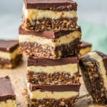 Layered dessert bars stacked on top of each other on a background of parchment and white countertop. Several bars are arranged around the stack.