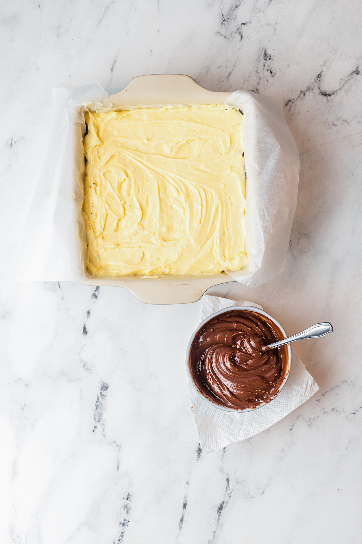 A baking dish with a layer of custard spread inside. Nearby is a bowl of melted chocolate.