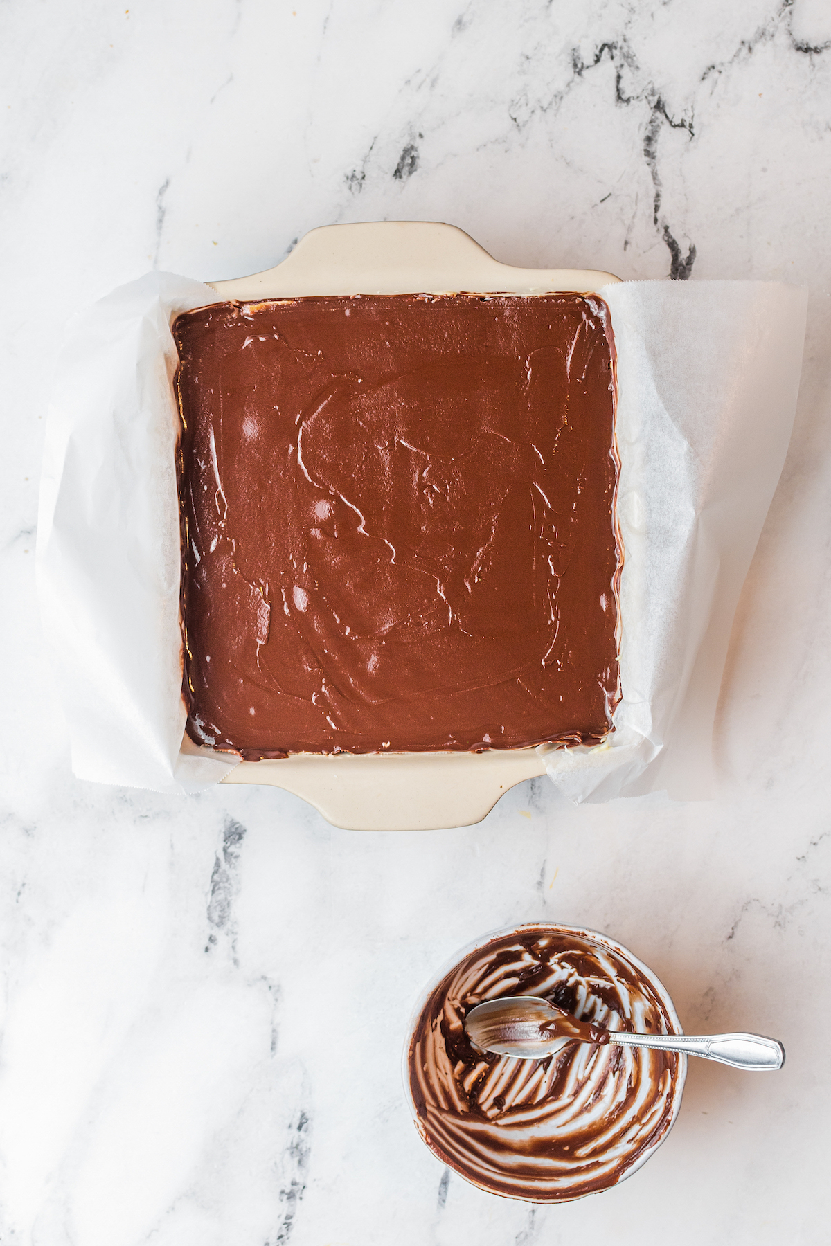 A square baking dish with a layer of melted chocolate topping visible inside. An empty, microwave-safe bowl with some chocolate still stuck to the sides sits on the countertop nearby.