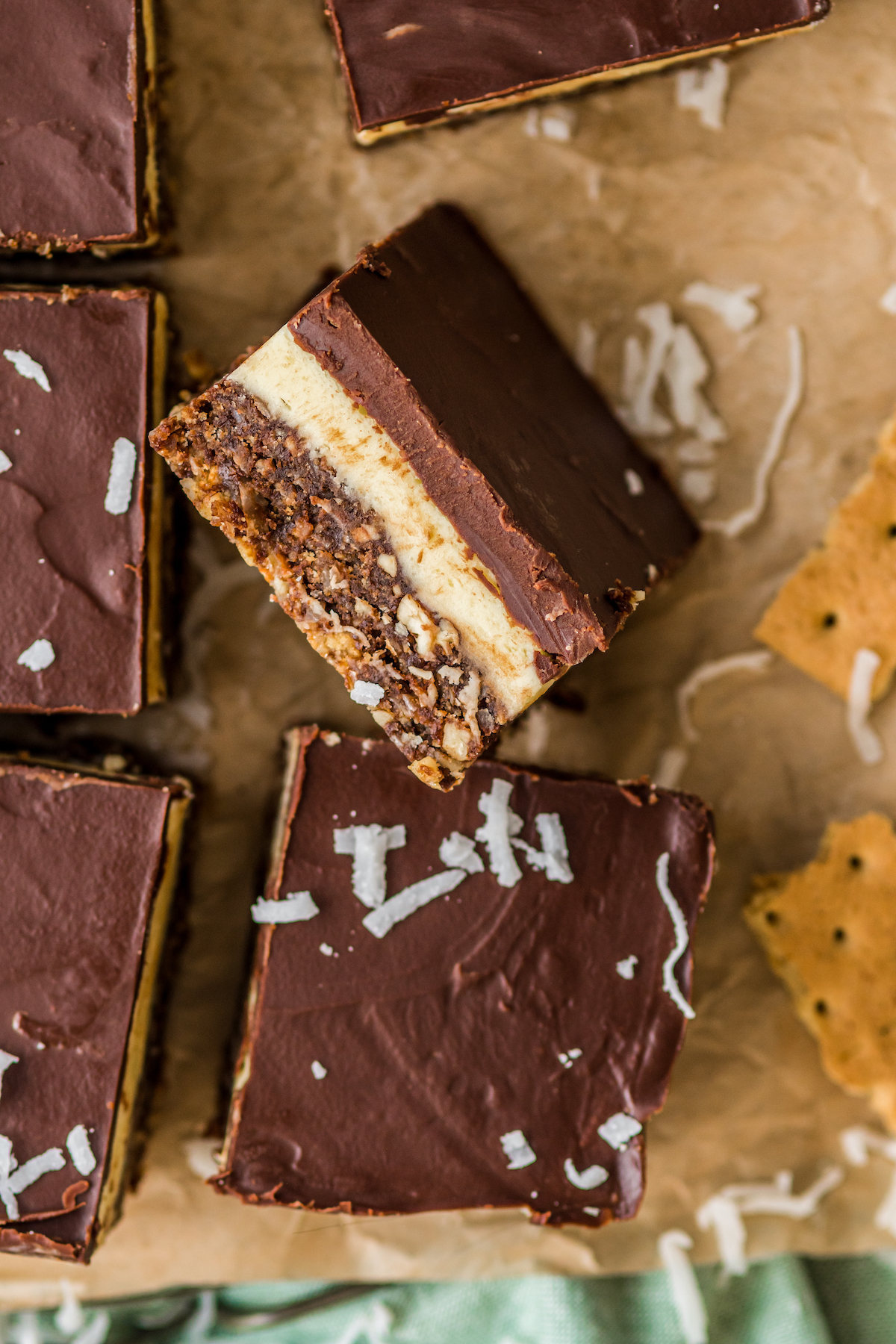 Several nanaimo bars on a sheet of parchment, decorated with shreds of coconut and a couple of broken graham crackers.