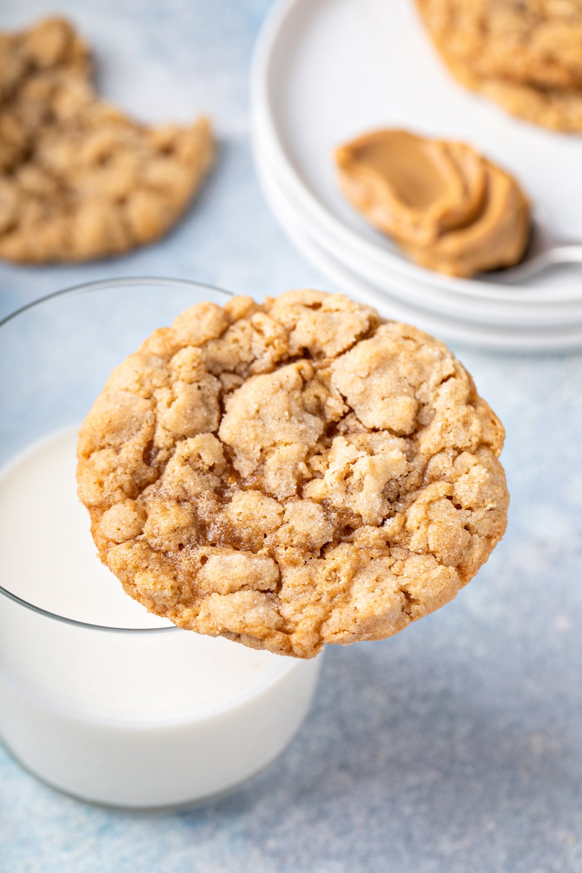 A peanut butter cookie balanced on the rim of a glass of milk. A saucer with pieces of cookie is in the background.