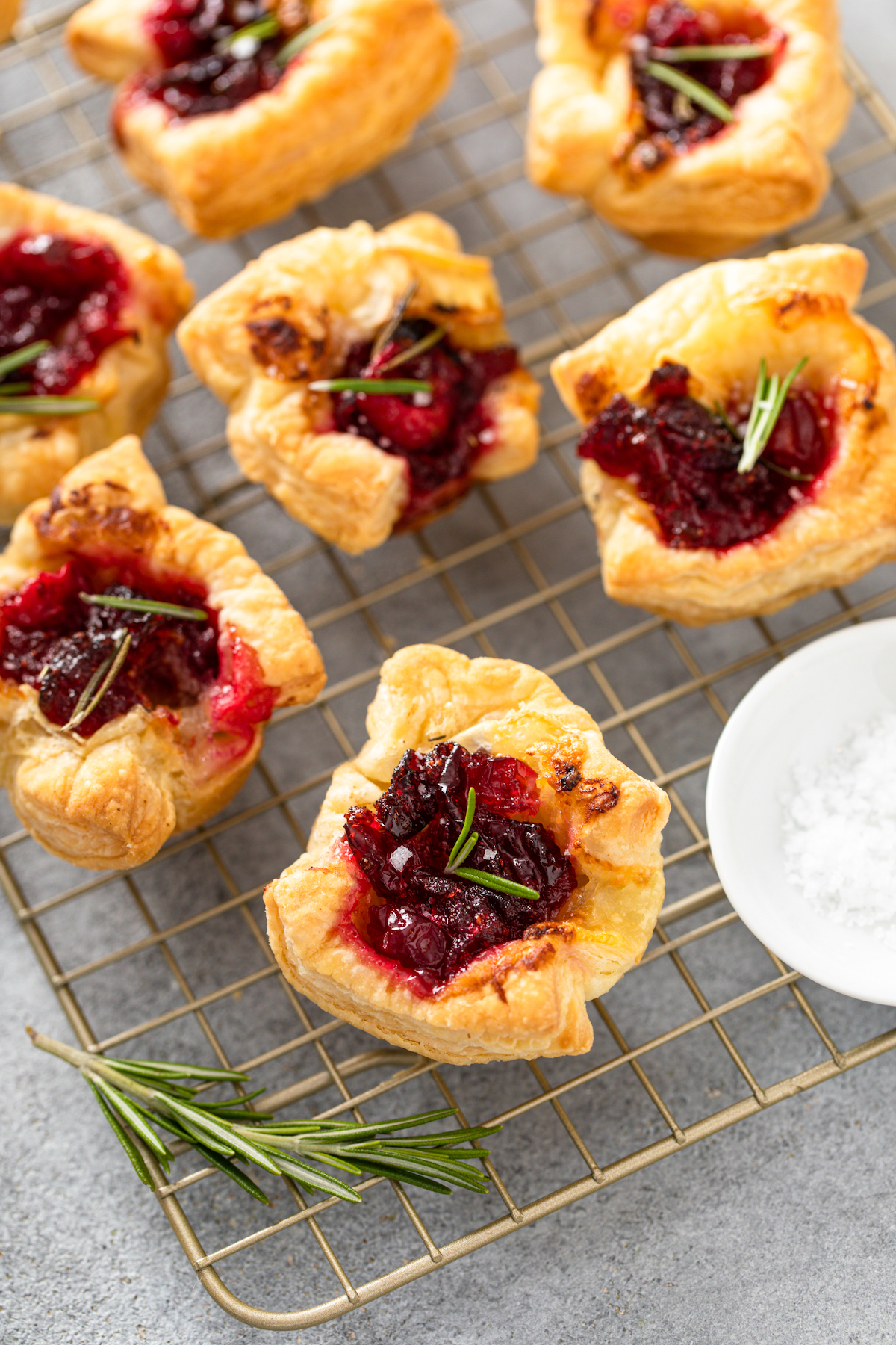 Cranberry Brie Bites on a cooling rack with fresh rosemary and flaky sea salt in a bowl.