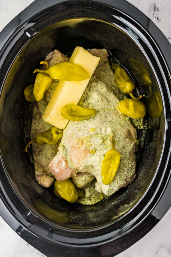 Overhead shot of a crockpot insert, filled with raw chicken, seasoning mix, a stick of butter, and pepperoncini.