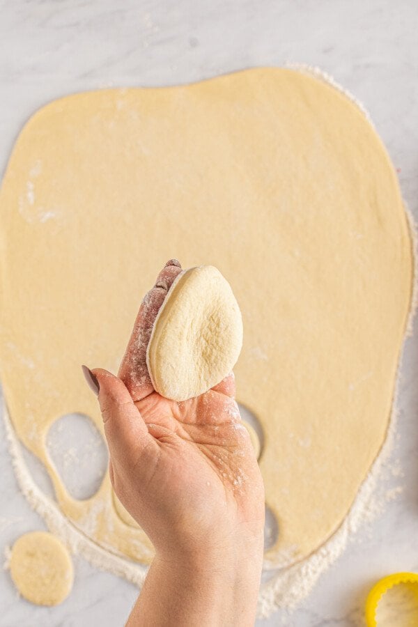 A circle of dough has been cut from the rolled-out donut dough. A hand is holding the circle of dough toward the cameral.